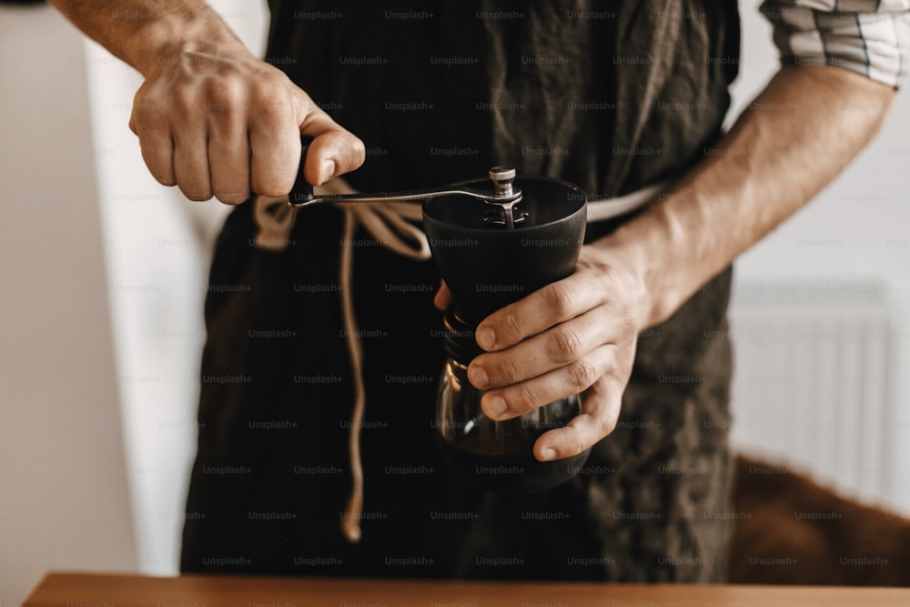 黒のスタイリッシュなエプロンでプロのバリスタがエアロプレス用のコーヒーを挽く、代替の醸造方法。コーヒー豆の手動グラインダーを持つ手。代替コーヒーのアイテム