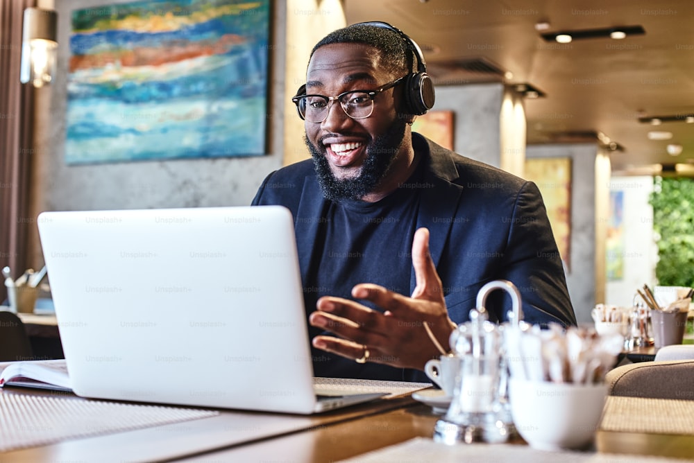 Positive stylish young Afro American businessman in dark-blue jacket and glasses sitting at the table, indoors, wearing wireless earbuds, chatting with his boss via video conference call on generic laptop computer, looking at screen and smiling cheerfully