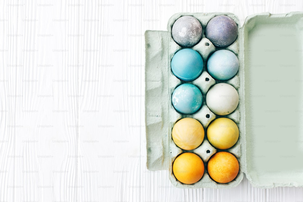 흰색 나무 배경에 판지 트레이에 있는 세련된 부활절 달걀, 텍스트를 위한 공간. 무지개 색상의 파스텔 천연 염료로 칠해진 현대적인 다채로운 부활절 달걀. 행복한 부활절. 휴일 장식