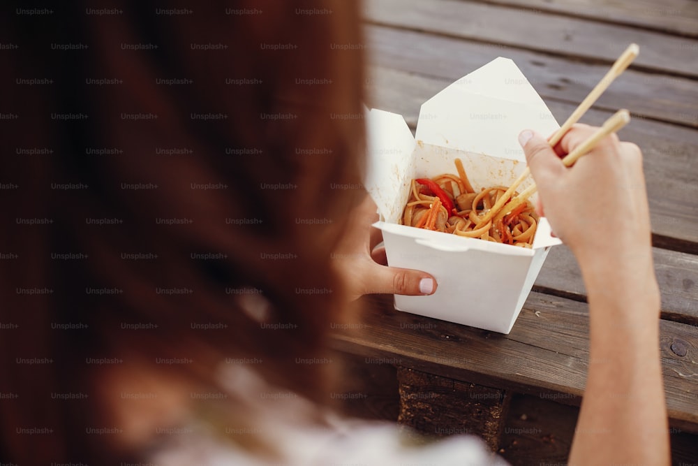 Wok with noodles and vegetables in carton box to go and bamboo chopsticks. Traditional Asian cuisine. Asian Street food festival. Girl eating thai noodles in open box takeaway. Food delivery