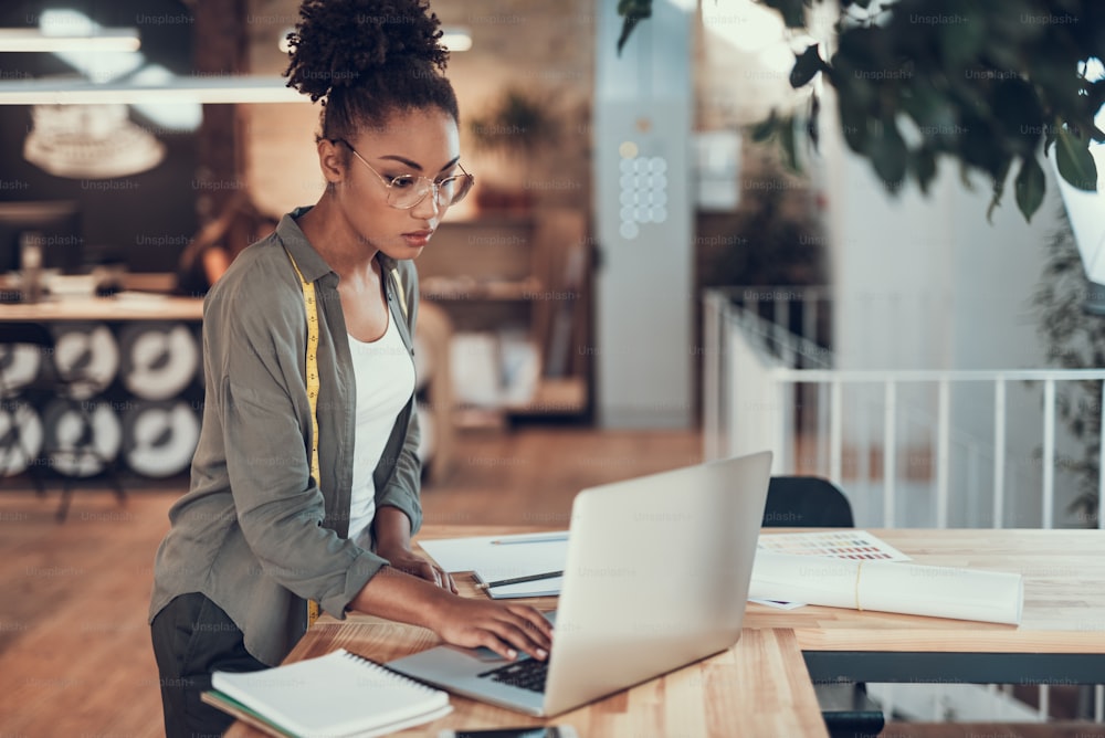 Portrait of beautiful young woman with measuring tape typing on notebook while standing near office desk with stationery. She is looking at display with serious expression