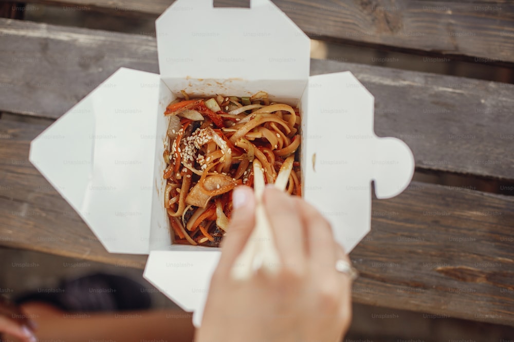 Wok with noodles and vegetables in carton box to go and bamboo chopsticks. Traditional Asian cuisine. Asian Street food festival. Girl eating thai noodles in open box takeaway. Food delivery