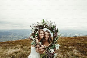stylish bridesmaid and gorgeous bride laughing and looking through wreath, boho wedding, luxury ceremony at mountains