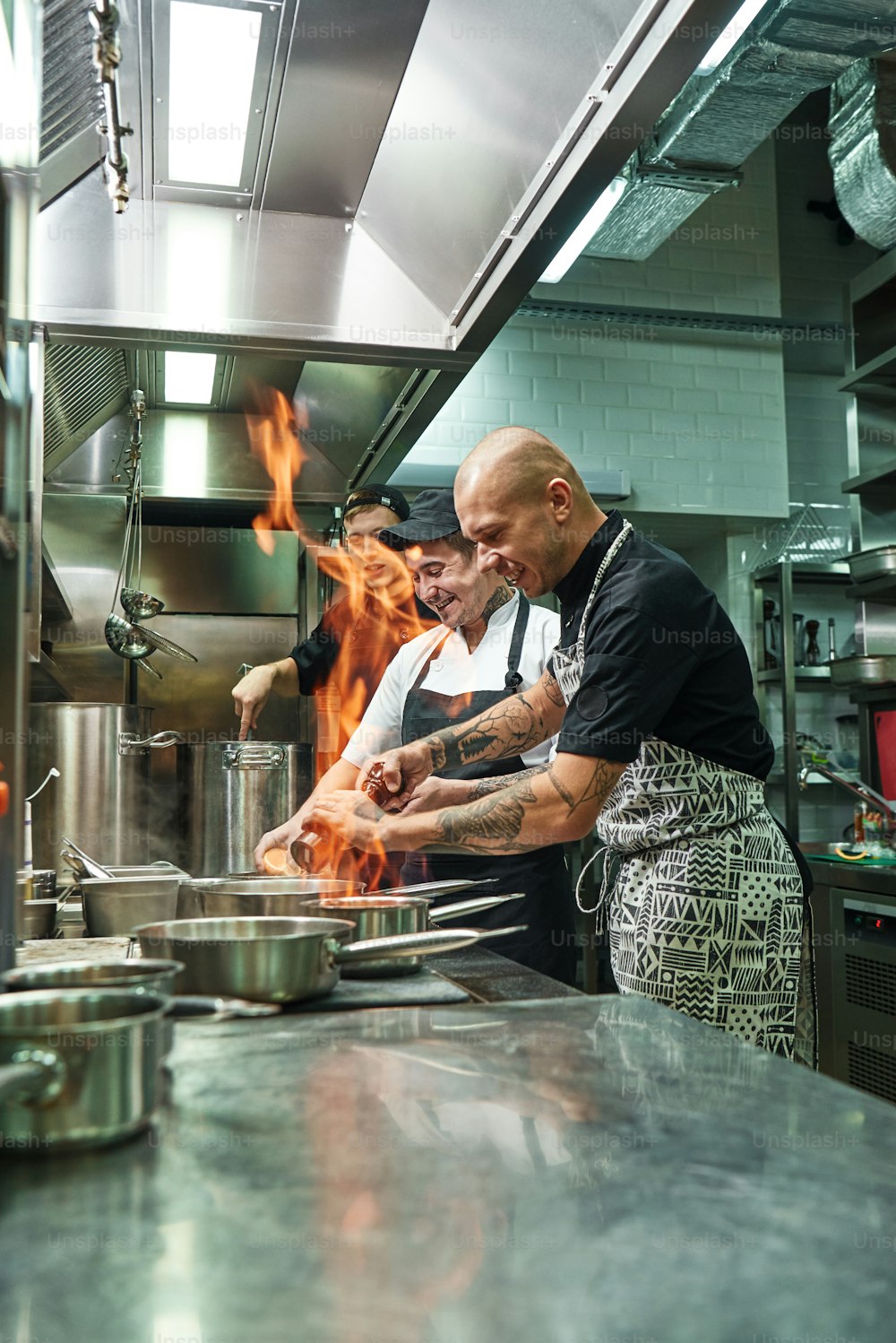 매우 뜨거운 쾌활한 요리사와 두 명의 조수가 레스토랑의 부엌에서 열린 불로 스토브에서 요리를 준비하고 있습니다. 플람베. 요리 개념