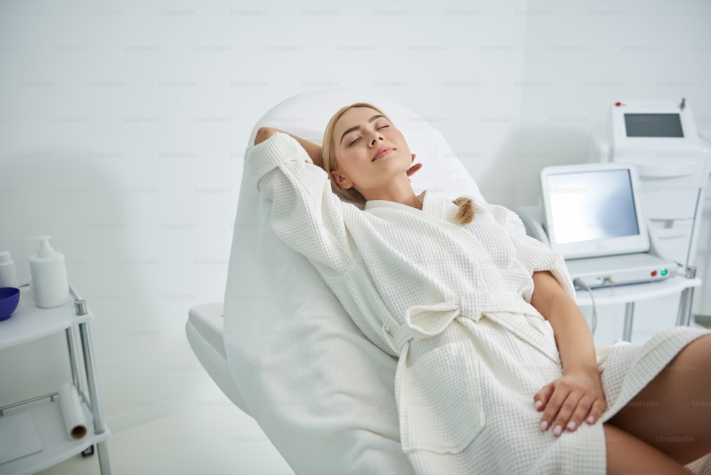 Relaxing at spa salon. Charming blond girl in white soft bathrobe lying on comfortable daybed and putting hand behind her head