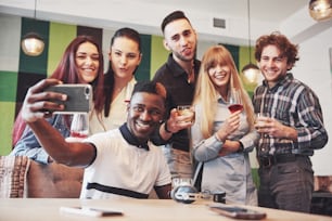 Friends having fun at restaurant. Three boys and three girls making selfie and laughing. On foreground boy holding smart phone. All wear casual clothes.