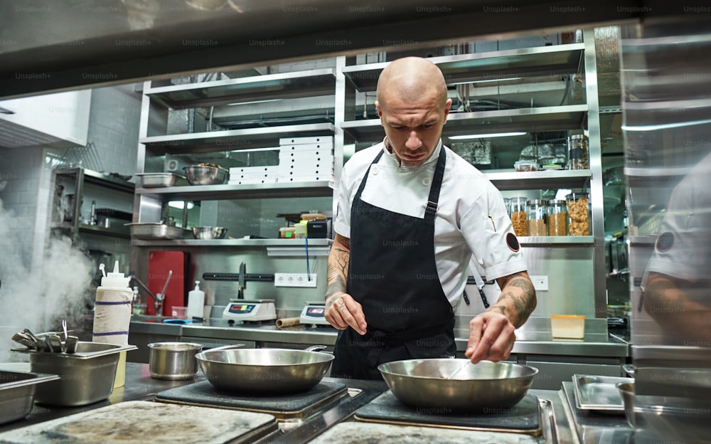 Confidence in everything. Serious male chef with several tattoos on his arms frying ingredients for his dish in a restaurant kitchen. Cooking process. Food blogger