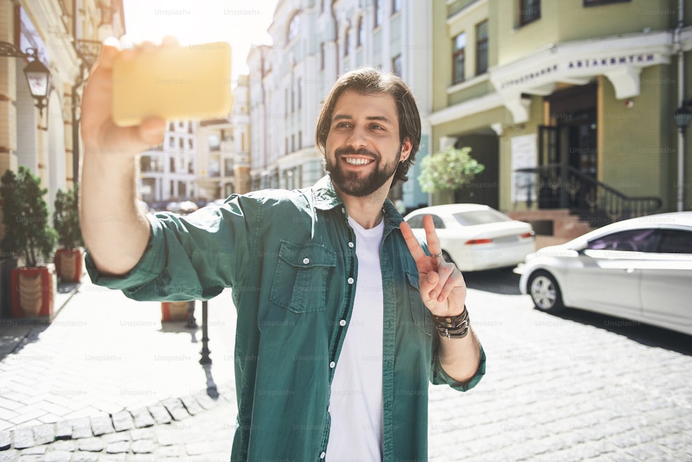 Para la memoria. Retrato de cintura para arriba de una persona masculina riendo de pie en la calle con un teléfono inteligente. Se está fotografiando a sí mismo y mostrando un gesto de paz ante la cámara