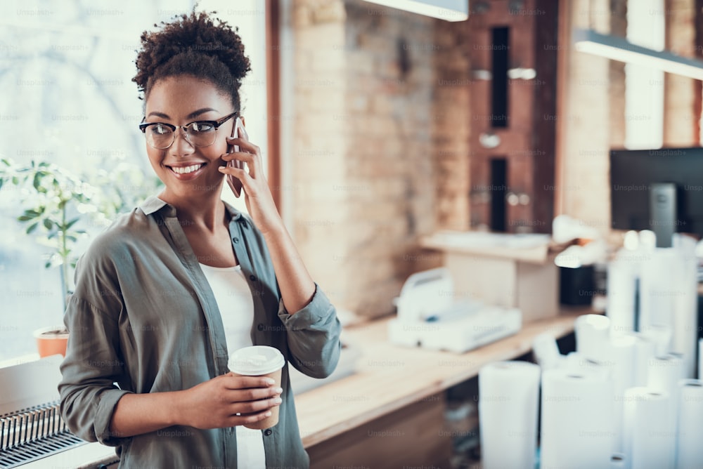 Waist up portrait of attractive young lady in glasses holding cup of coffee while having phone conversation. She is looking away and smiling