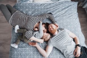 Top view of beautiful young mother, father and their daughter looking at camera and smiling while lying on bed head to head.