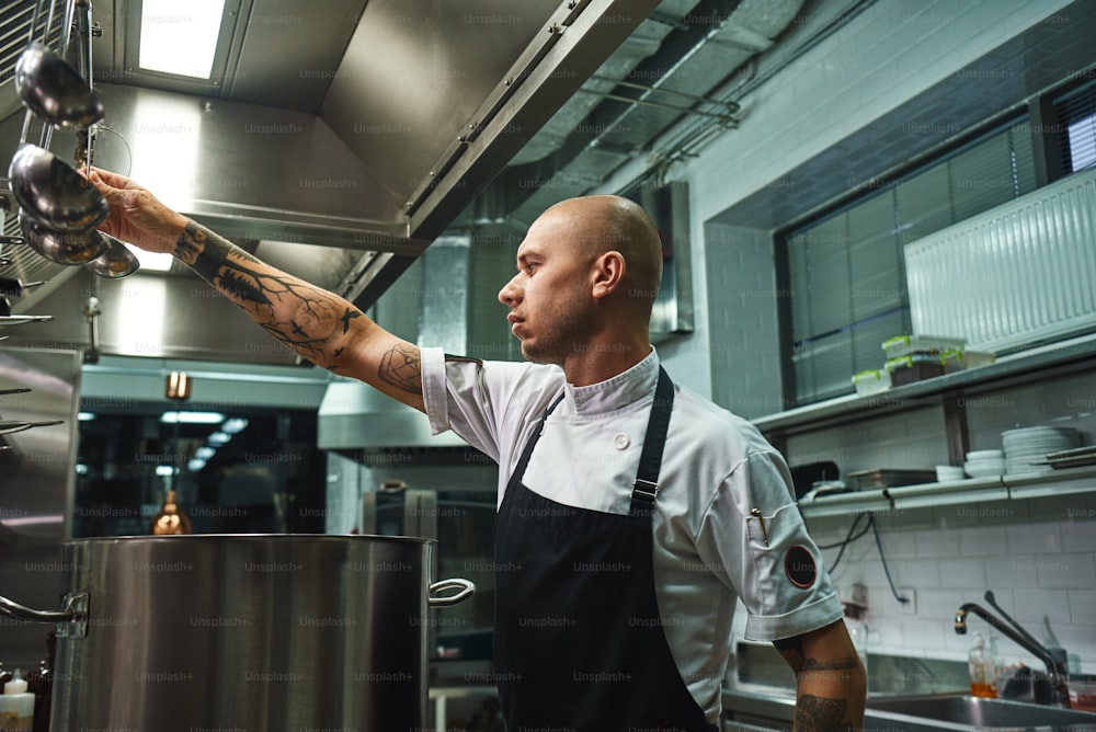 Famous soup. SIde view of attractive bald chef in apron taking a ladle for soup cooking while standing in a restaurant kitchen. Cooking concept