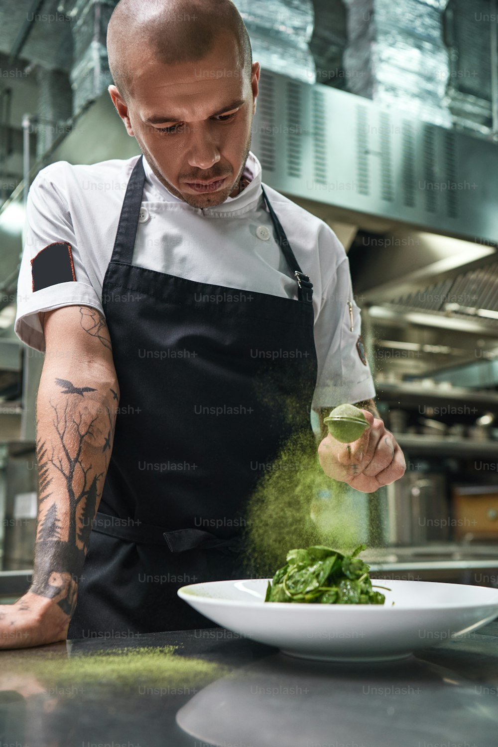 Alimentación saludable. Retrato vertical de un guapo chef masculino con delantal negro agregando especias en ensalada mientras está de pie en la cocina de un restaurante.
