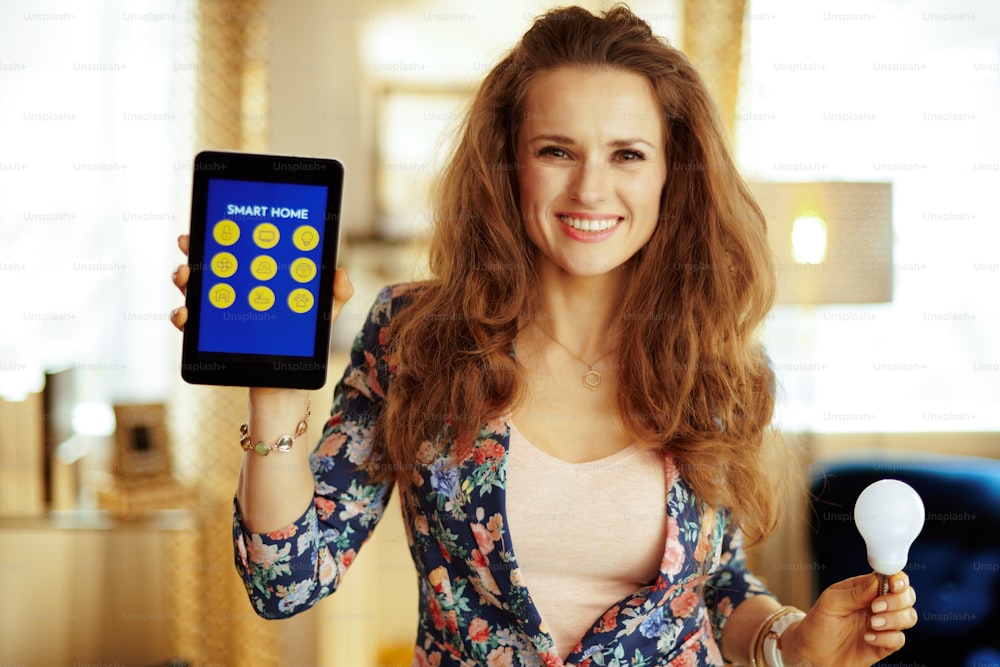Retrato de una joven ama de casa sonriente con cabello largo y castaño que muestra una tableta PC con una aplicación para el hogar inteligente y una lámpara inteligente en la sala de estar moderna.