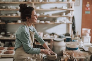 Side view portrait of beautiful craftswoman making clay bowl in pottery workshop. She looking away and smiling