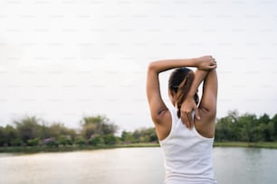 Healthy young Asian runner woman warm up the body stretching before exercise and yoga near lake at park under warm light morning. Lifestyle fitness and active women exercise in urban city concept.