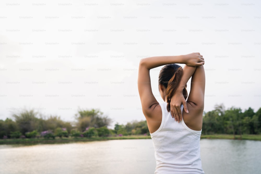 La joven corredora asiática saludable calienta el estiramiento del cuerpo antes del ejercicio y el yoga cerca del lago en el parque bajo una mañana de luz cálida. El estilo de vida de la aptitud y el ejercicio de las mujeres activas en el concepto de la ciudad urbana.