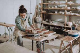 Portrait of charming craftswoman sitting on bench while kneading and shaping clay in pottery workshop. She looking down with serious expression