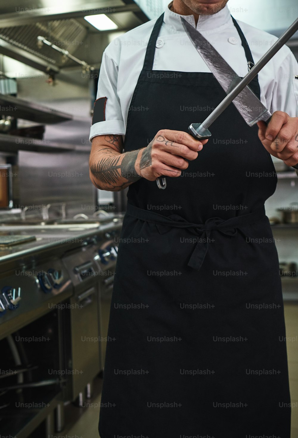 Pronto da cuocere. Foto verticale di giovane in grembiule con tatuaggi sulle braccia che affila un coltello nella cucina di un ristorante. Concetto di cottura