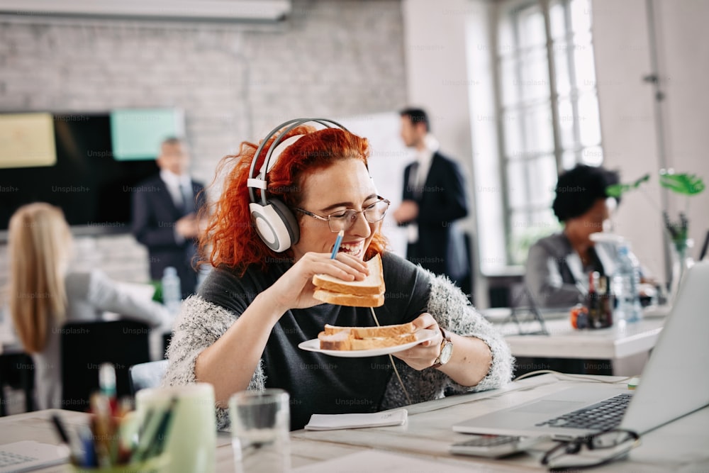 Happy casual businesswoman having fun and laughing while eating sandwich on a break at work. There are people in the background.