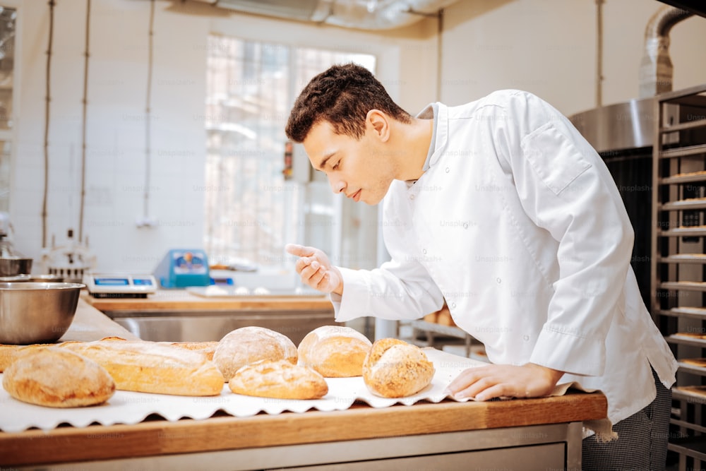 Just baked bread. Dark-haired baker wearing white jacket feeling satisfied while smelling just baked bread