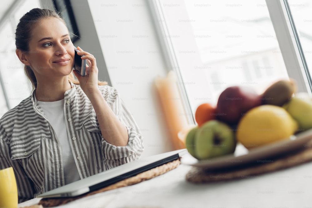 Actividad freelance en casa. Retrato de cintura para arriba de joven dama sonriente hablando por teléfono inteligente mientras está sentada en la mesa con computadora portátil y frutas en el plato