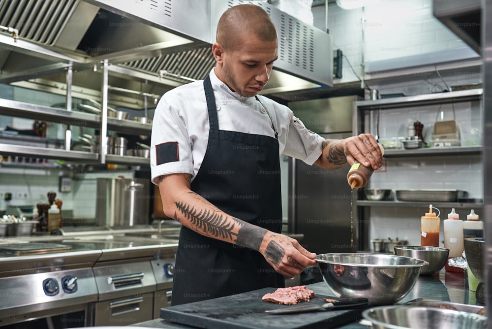 Câmera lenta. Bonito chef de restaurante em avental e com tatuagens nos braços adicionando seu famoso molho ao prato. Processo de cozimento