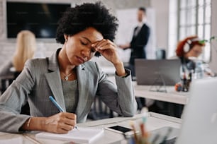 Imprenditrice afroamericana che fa un piano e scrive appunti mentre lavora alla sua scrivania in ufficio. Ci sono persone sullo sfondo.