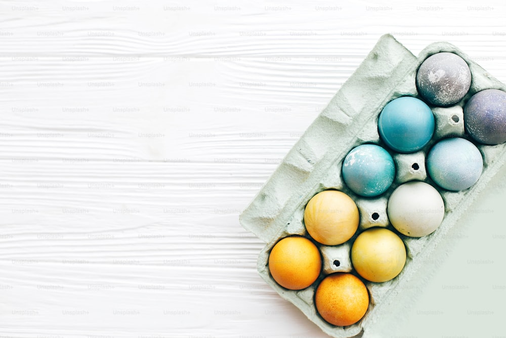 행복한 부활절. 흰색 나무 배경에 판지 트레이에 무지개 파스텔 색상의 세련된 부활절 달걀, 텍스트를 위한 공간이 있는 평평한 누워. 천연 염료로 칠해진 현대적인 다채로운 부활절 달걀