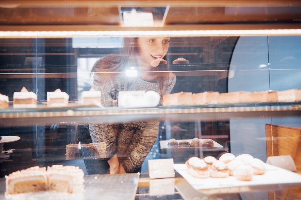 Mulher sorridente na câmera através da vitrine com doces e bolos no interior do café moderno.
