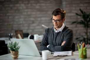 Satisfied entrepreneur with arms crossed reading good news on a computer while working in the office.