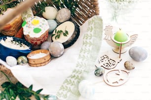 Happy Easter. Stylish Easter eggs, easter bread cake, ham, beets, sausage, butter, green branches in wicker basket on white wooden background with easter candle and decorations