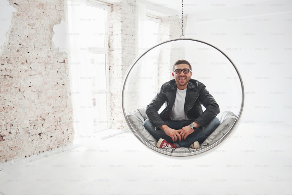 Full-length portrait of relaxed hipster man in casual sitting on hanging chair bullet and smiling on camera isolated over white background.