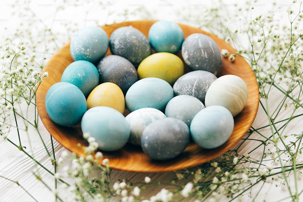 Felices Pascuas. Elegantes huevos de Pascua con flores de primavera en plato de madera sobre fondo de madera blanco. Huevos de pascua modernos pintados con tinte natural en colores mármol amarillo, azul, verde, gris