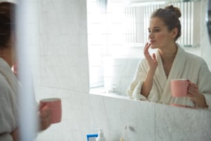セルフケアのコンセプト。白いバスローブを着た若い笑顔の女性がコーヒーを飲みながら朝を楽しみ、鏡の前で唇に優しく触れている腰の上の反射