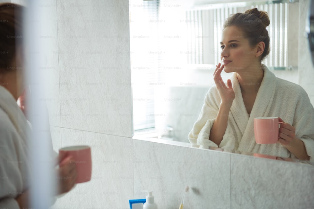 Concepto de autocuidado. Reflejo de cintura para arriba de una joven mujer sonriente en bata de baño blanca disfrutando de su mañana con una taza de café y tocando tiernamente sus labios ante el espejo