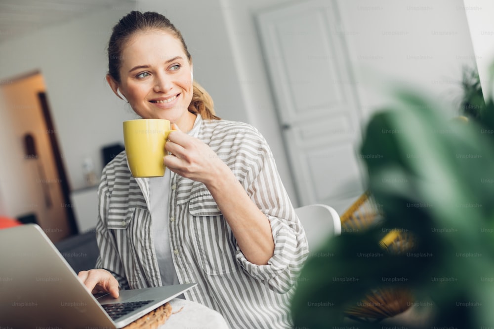 Freelance activity. Waist up portrait of happy smiling lady sitting on table with laptop on it and drinking coffee. Home interior around