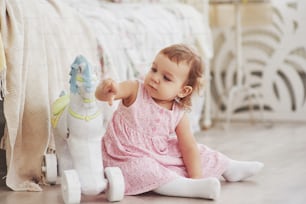 Niña en lindo vestido sentada en la cama jugando con juguetes en el hogar. Habitación infantil vintage blanca. Concepto de infancia.