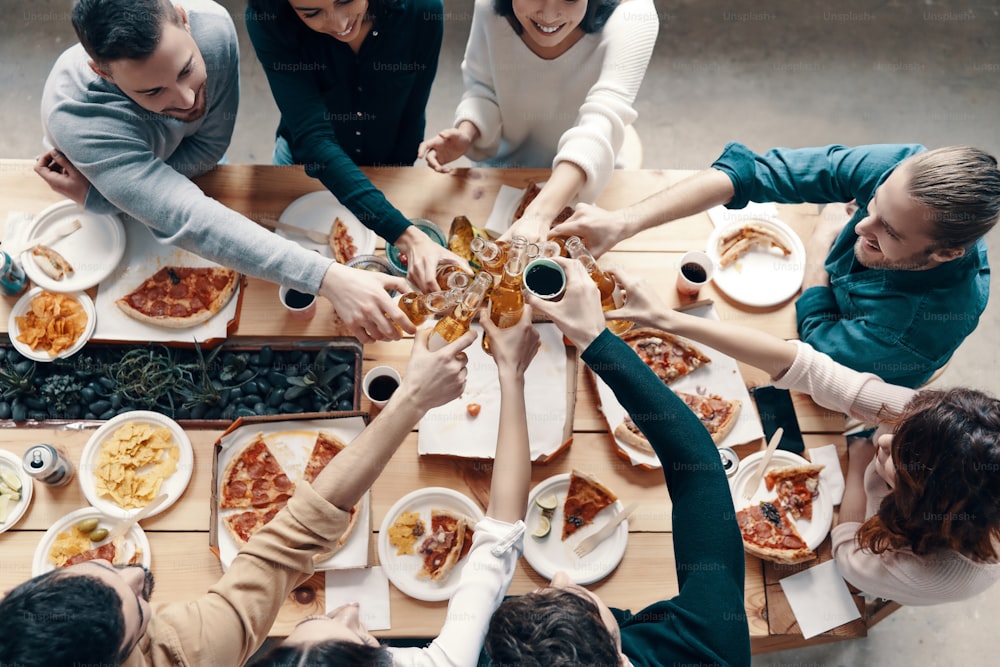 Top view of young people in casual wear toasting each other and smiling while having a dinner party indoors