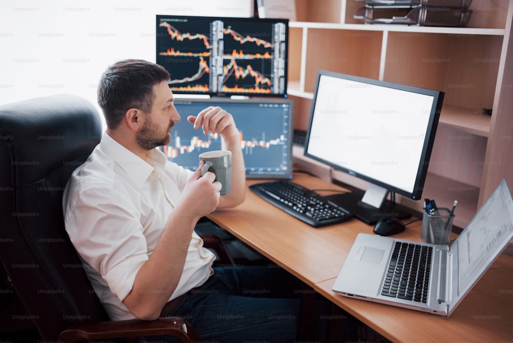 Young businessman is sitting in office at table, working on computer with many monitors,diagrams on monitor. Stock broker analyzes binary options charts.Hipster man drinking coffee,studying.