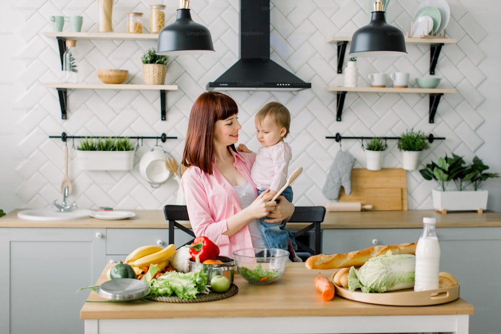 Cheerful beautiful mother in a pink shirt is preparing a fresh vegetable salad at home in the kitchen, holding her little cute daughter. Mother and daughter, healthy food concept