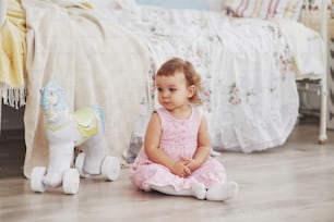 Baby girl in cute dress siting at bed playing with toys by the home. White vintage childroom. Childhood concept.