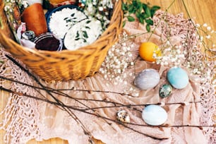Stylish Easter basket with modern eggs, easter bread cake, ham, beets, sausage, butter and green branches on rustic fabric with spring flowers in sunny light. Happy Easter. Food for holiday