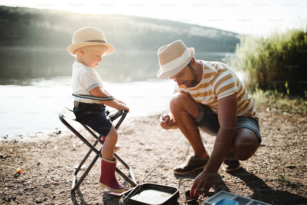 A mature father with a small toddler son outdoors fishing by a river or a lake, talking.