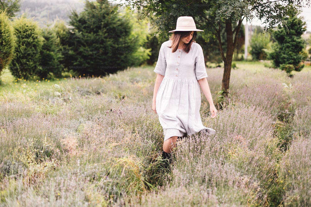 Stylish hipster girl in linen dress and hat walking in lavender field and smiling. Happy bohemian woman relaxing and enjoying lavender aroma. Atmospheric calm rural moment. Space for text