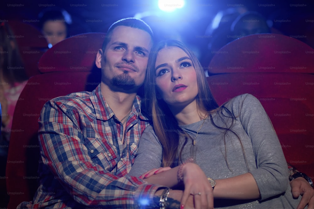 Vista frontale della coppia seduta insieme al cinema, guardando una commedia o un film romantico. Bel uomo barbuto che abbraccia la bella fidanzata seduta vicino mentre recensisce il film. Concetto di tempo libero.