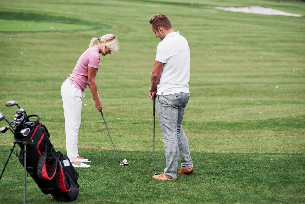 Adult man teaching the woman how to play the golf on the field with green grass.