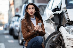 Young unfortunate woman using smart phone and text messaging after a car accident in the city.