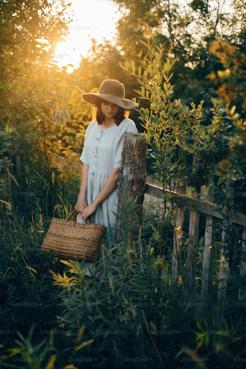 Stylish girl in linen dress holding rustic straw basket at wooden fence  in sunset light. Boho woman relaxing and posing in summer countryside in warm evening. Atmospheric rural moment