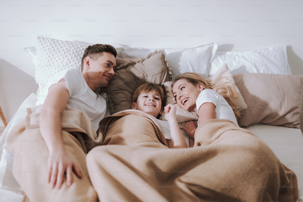 Pleasant morning. Cheerful parents lying on soft pillows in bed and their son smiling while relaxing with them