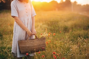 Stylish girl in linen dress holding rustic straw basket with poppy flowers in meadow in sunset light. Boho woman relaxing and gathering wildflowers in summer field. Atmospheric moment